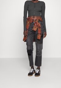 Grå ribbad croptopp med knappstängning, parat med svarta patchworkjeans med olika texturer och en röd rutigt flanellskjorta knuten i midjan.