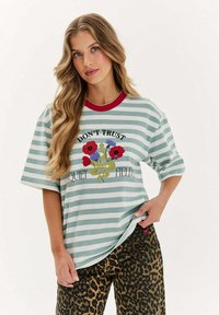 Pruhované tričko v světle modré a bílé barvě s červeným límcem, s květinovou grafikou a nápisem. V kombinaci s kalhotami s leopardím vzorem.