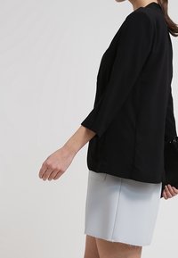 Kobieta w czarnym żakiecie i jasnoszarej spódnicy, trzymająca małą czarną torebkę i z włosami związanymi do tyłu.