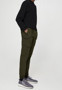 Zwarte gebreide trui, olijfgroene cargobroek met zakken en grijze sneakers met een gestructureerde afwerking en witte zolen. Kettingdetail aan de broek.