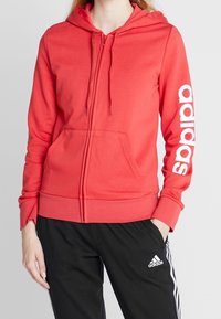 Sweat à capuche rouge zippé avec une poche kangourou et le logo "adidas" blanc sur la manche gauche. Tissu doux avec une coupe décontractée et une capuche ajustable.