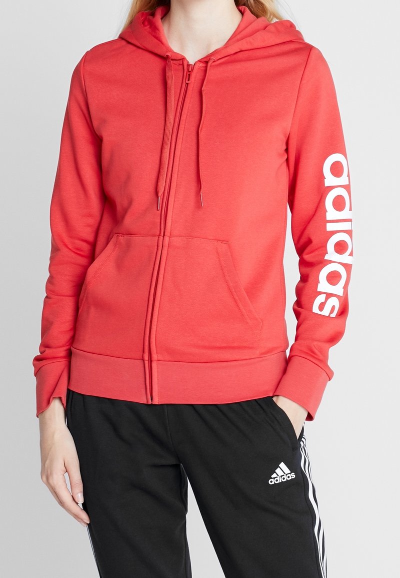 Sweat à capuche rouge zippé avec une poche kangourou et le logo "adidas" blanc sur la manche gauche. Tissu doux avec une coupe décontractée et une capuche ajustable.