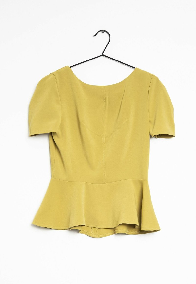 Blouse jaune à manches courtes avec basque suspendue sur un cintre métallique noir sur fond blanc.