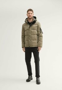 Gepolsterte olivgrüne Jacke mit Kapuze, mit einer Reißverschlusstasche auf der Brust, kontrastierenden schwarzen Akzenten und einer strukturierten Oberfläche.