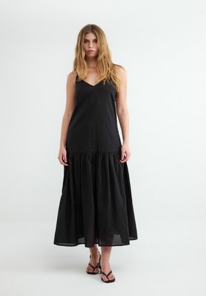 Kobieta o długich blond włosach, ubrana w czarną sukienkę maxi bez rękawów i czarne sandały na paski, stojąca na białym tle.