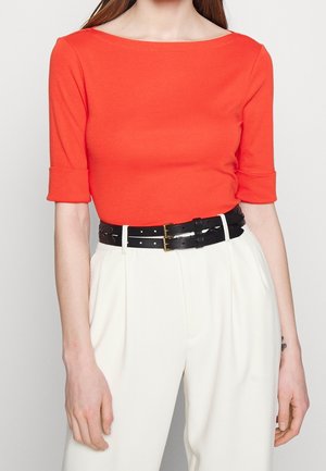 Femme portant un haut près du corps orange à manches aux coudes, rentré dans un pantalon blanc taille haute avec une ceinture noire à double boucle.