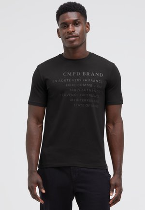Young man wearing a black CMPD BRAND t-shirt with French and English text standing against a plain light background.