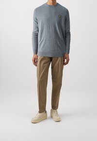 Maglione azzurro chiaro con orlo e polsini a coste, che presenta un piccolo logo ricamato. Abbinato a pantaloni beige e sneakers chiare.