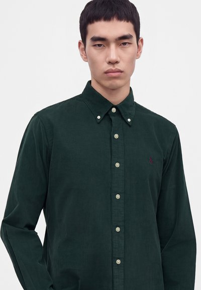 Jeune homme aux cheveux courts noirs portant une chemise verte foncée à boutons avec un petit logo brodé sur la poitrine.