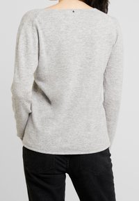 Grauer Langarmpullover mit leicht offenem Rücken, strukturiertem Strick und einem kleinen Logodetail am Halsausschnitt. Kombiniert mit schwarzen Hosen.