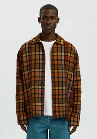 Bruine en oranje geruite blouse van flanel, met knopen en een ruime kraag, losse pasvorm, gedragen over een wit T-shirt, gecombineerd met een blauwe spijkerbroek.