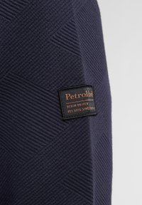 Niebieska tkanina teksturowana z geometrycznym wzorem; zawiera czarną metkę z pomarańczowym napisem "PetrolInd" oraz "DENIM DRIVEN."