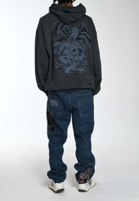Felpa nera con cappuccio decorata con un grande design grafico blu, abbinata a jeans blu scuri con dettagli ricamati di una pantera e di un drago. Sneakers bianche.