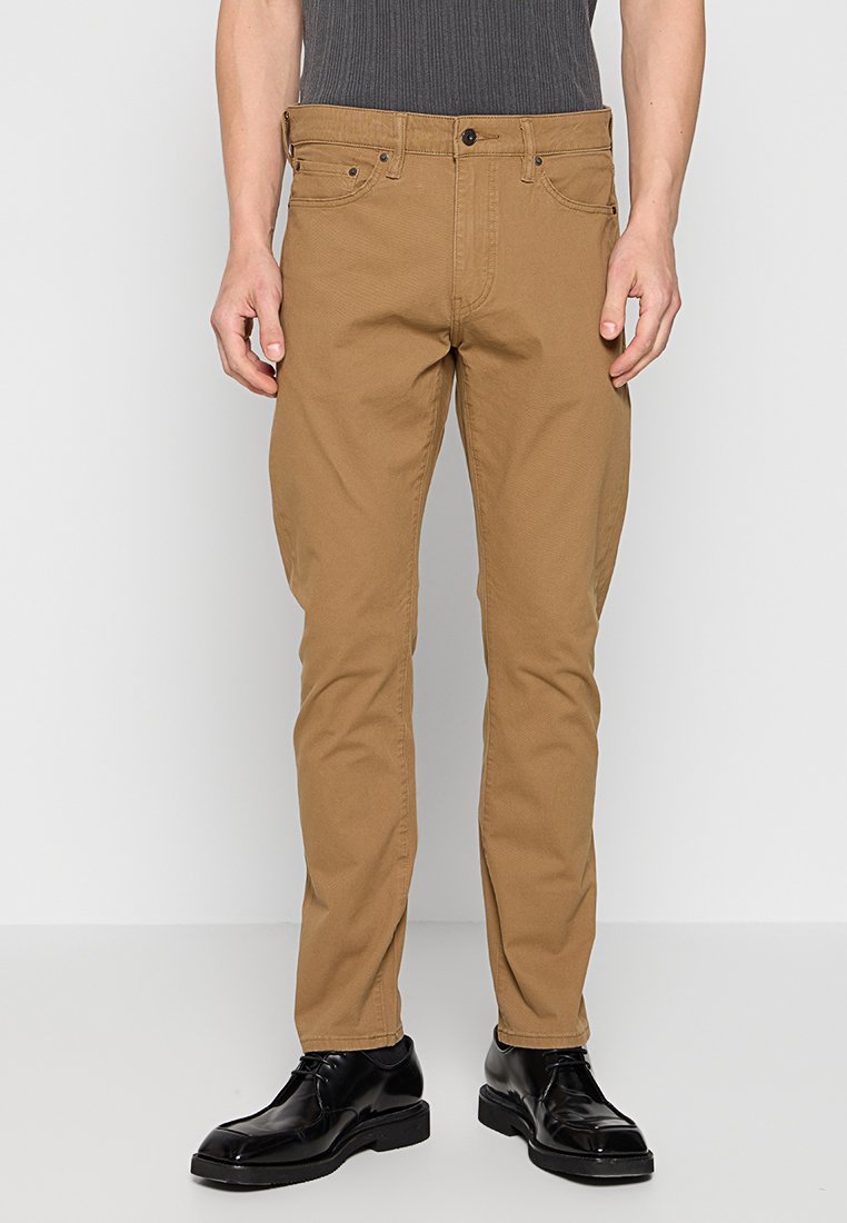 Tan katoenen broek met een recht model, voorzien van voorkeurszakken, een knoopsluiting en ton-sur-ton stiksels. Gewerkt met zwarte schoenen.