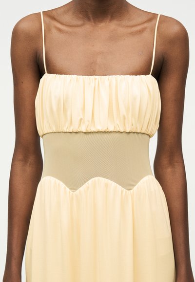 Γυναίκα που φορά αμάνικο, ανοιχτό κίτρινο φόρεμα με ραφτό μπούστο, λεπτές τιράντες και εφαρμοστό σχέδιο στη μέση, μπροστά από απλό φόντο.