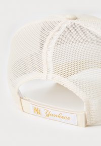 Hvid mesh baseballcap med en flad skygge, justerbar rem og et guld "Yankees" logomerke på bagsiden. Struktureret materiale, afslappet design.
