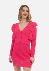 Rosa stickad cardigan med volangdetaljer, knäppning och V-ringning, i kombination med en matchande texturerad kjol. Båda är gjorda av mjukt tyg.