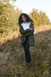 Padded olive green coat with a hood over a white shirt, paired with black pants, set in a grassy, natural outdoor environment.