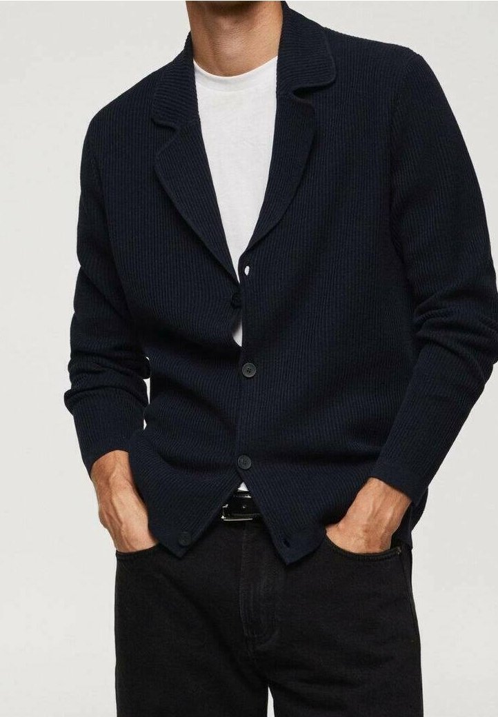 Homme portant un cardigan côtelé foncé par-dessus un t-shirt blanc, les mains dans les poches d’un pantalon noir, debout devant un fond uni.