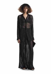 Schwarz, transparente, plissierte lange Bluse mit durchgehenden Knöpfen, im lässigen Schnitt mit seitlichen Schlitzen, getragen über einem passenden Rock.