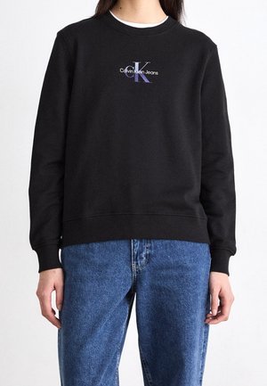 Musta pitkähihainen Calvin Klein Jeans -collegepaita, jossa rintakehän logo, sinisten farkkujen kanssa, kuvattuna olkapäistä lantioihin.