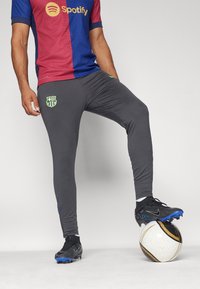 Grijze sportbroek met een gladde textuur, voorzien van een groen Barcelona-logo en blauwe accenten, gecombineerd met zwarte voetbalschoenen en een gemarmerde bal.