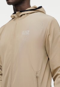 Άνδρας που φοράει μπεζ μπουφάν με κουκούλα, εμπρός φερμουάρ και λευκό λογότυπο EA7 Emporio Armani στο στήθος.