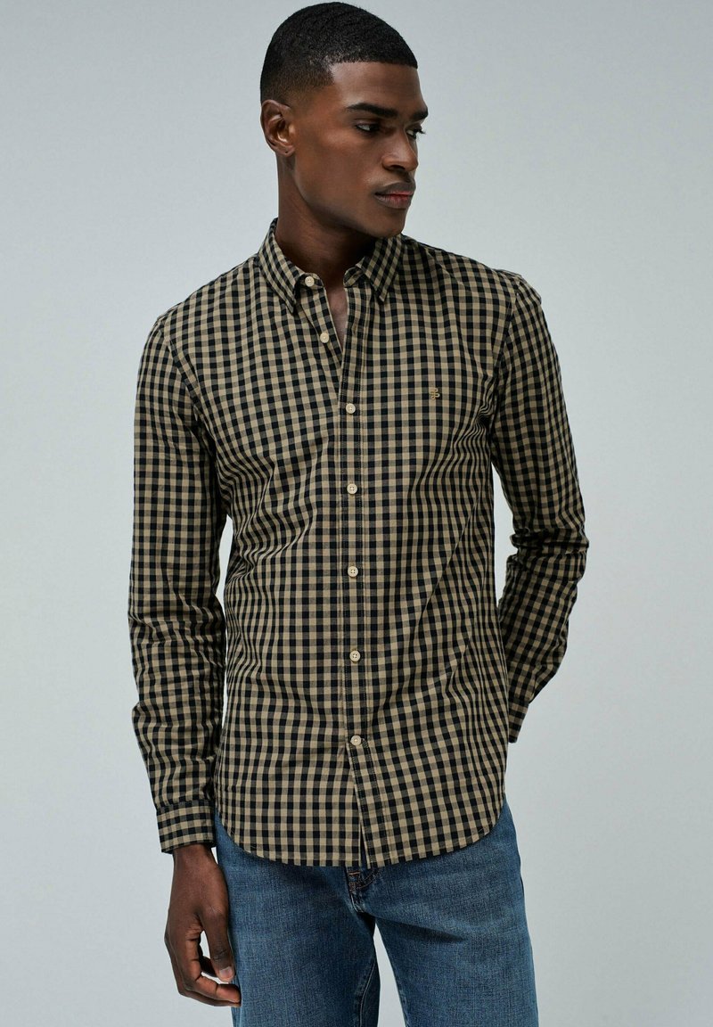 Camisa abotonada en patrón de cuadros negro y beige, con mangas largas, cuello y un sutil detalle de logo en el pecho. Usada con jeans azules.