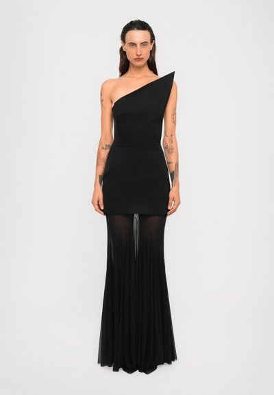 Model with tattoos wears a black one-shoulder dress with sheer, pleated floor-length skirt standing against a plain white background.