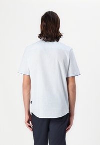 Camisa de manga curta azul claro com uma textura subtil, hem arredondado e costura contrastante. Apresenta um pequeno logotipo na parte inferior das costas.