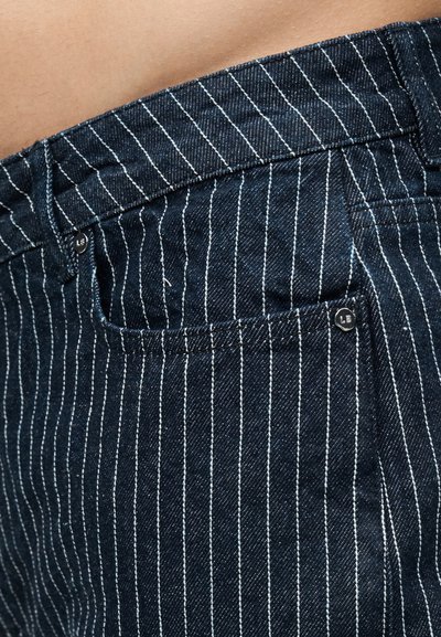 Κοντινό πλάνο σκούρου μπλε τζιν παντελονιού με λεπτές ρίγες, με ορατή μέση, θηλιά ζώνης και λεπτομέρειες ραφής στην μπροστινή τσέπη.