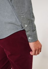Szara longsleeve w miękkim materiale, z mankietami zapinanymi na guziki i spiczastym wzorem rękawów, w połączeniu z ciemnogranatowymi spodniami.