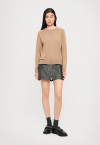 Asiatisk kvinne som står, iført en beige genser med et lite rosa hjerte, svart skinn mini-skjørt med glidelås foran, grå sokker og svarte loafers.