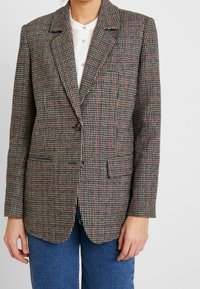 Hnědo-krémový blazer s houndstooth vzorem, s jedním knoflíkem, klopovými límci a dvěma předními kapsami, vyrobený z texturovaného materiálu.