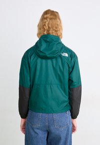 Roheline ja must jakk kapuutsiga, millel on sile tekstuur, elastsed mansetid ja logo seljal. Kantud kõrge vöökohaga teksadega.