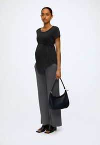 Mujer embarazada de pie, lleva una blusa negra con la cintura atada, pantalones grises de pierna ancha, tacones negros abiertos y sostiene un bolso negro.