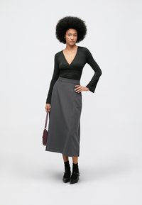 Top negro de manga larga con diseño cruzado, combinado con una falda gris hasta el suelo. La modelo sostiene un pequeño bolso burdeos y lleva zapatos negros.