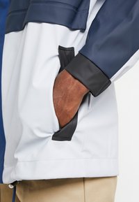 Main dans la poche d'une veste bleue et blanche avec bordure noire, portée sur un pantalon beige.