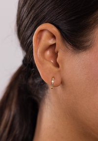 Goldene Creolen mit fünf eingefassten klaren Kristallen, einer glatten Textur und minimalistischem Design, vor einem hautfarbenen Hintergrund.