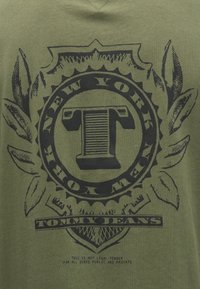 Roheline puuvillane T-särk, millel on must graafiline disain, sealhulgas ümmargune embleem, millel on tekst "NEW YORK" ja "TOMMY JEANS" ning leheaktsendid.