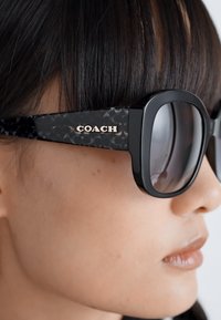 Ochelari de soare negri cu rame rectangulare supradimensionate, cu un model texturat pe brațe și cu logoul "COACH" în argintiu pe lateral.
