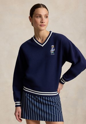Kobieta ubrana w granatowy sweter z emblematem niedźwiedzia i pasiastą granatową mini spódniczkę, stojąca z jedną ręką na biodrze.