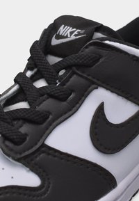 Στενή λήψη από μαύρο και άσπρο αθλητικό παπούτσι Nike με μαύρα κορδόνια, ραμμένο λογότυπο Swoosh της Nike και λογότυπο Nike στη γλώσσα, πάνω σε μονόχρωμο φόντο.