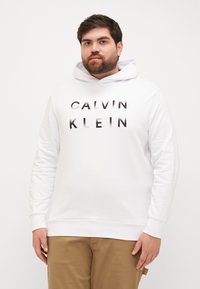 Biała bluza z kapturem wykonana z bawełnianego materiału, z przednią kieszenią typu kangura, ściągaczami przy mankietach oraz logo "CALVIN KLEIN" w czarnym napisie.