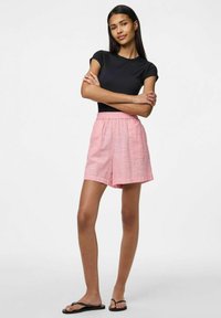 Rožinės patikrintos šortai su elastinga juosmena, derinami su juodu aptemptu marškinėliu. Modelis dėvi juodus šlepetes. Paprastas, kasdienis dizainas.