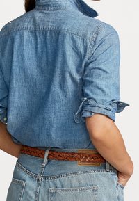 Ljusblå denimskjorta med uppvikta ärmar, med krage och ryggplatta. Bärs med ett flätat brunt läderbälte över ljusblå jeans.