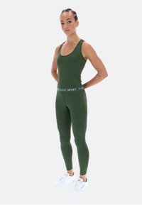 Zöld sportos trikó és leggings fehér logós derékrésszel. A modell fehér tornacipőt visel, és egy sima háttér előtt áll.