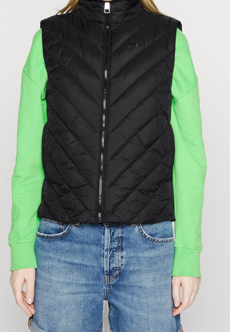 Black, quilted sleeveless vest with chevron pattern, front zipper, and small logo. Layered over bright green long-sleeve shirt.