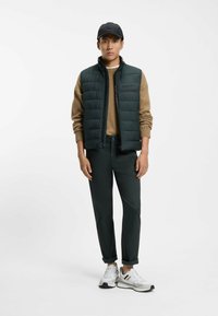 Gilet imbottito verde scuro sopra un maglione color cammello, abbinato a pantaloni verde scuro e sneakers bianche. Accessori includono un cappello nero.