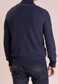 Mörkblå stickad tröja med hög krage och ribbade ärmslut, med ett texturerat mönster och kroppsnära passform. Bakifrån visar den axelsömmarna.